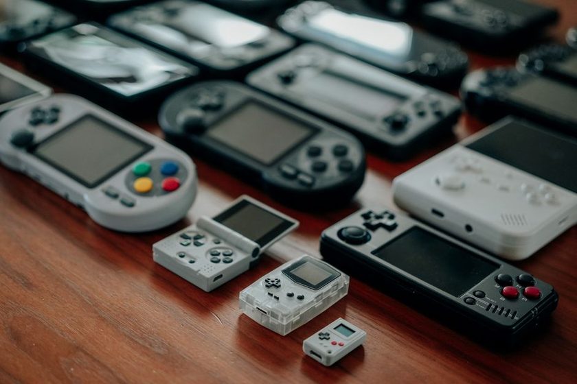 Três consoles portáteis de jogos lado a lado sobre mesa de madeira para comparação